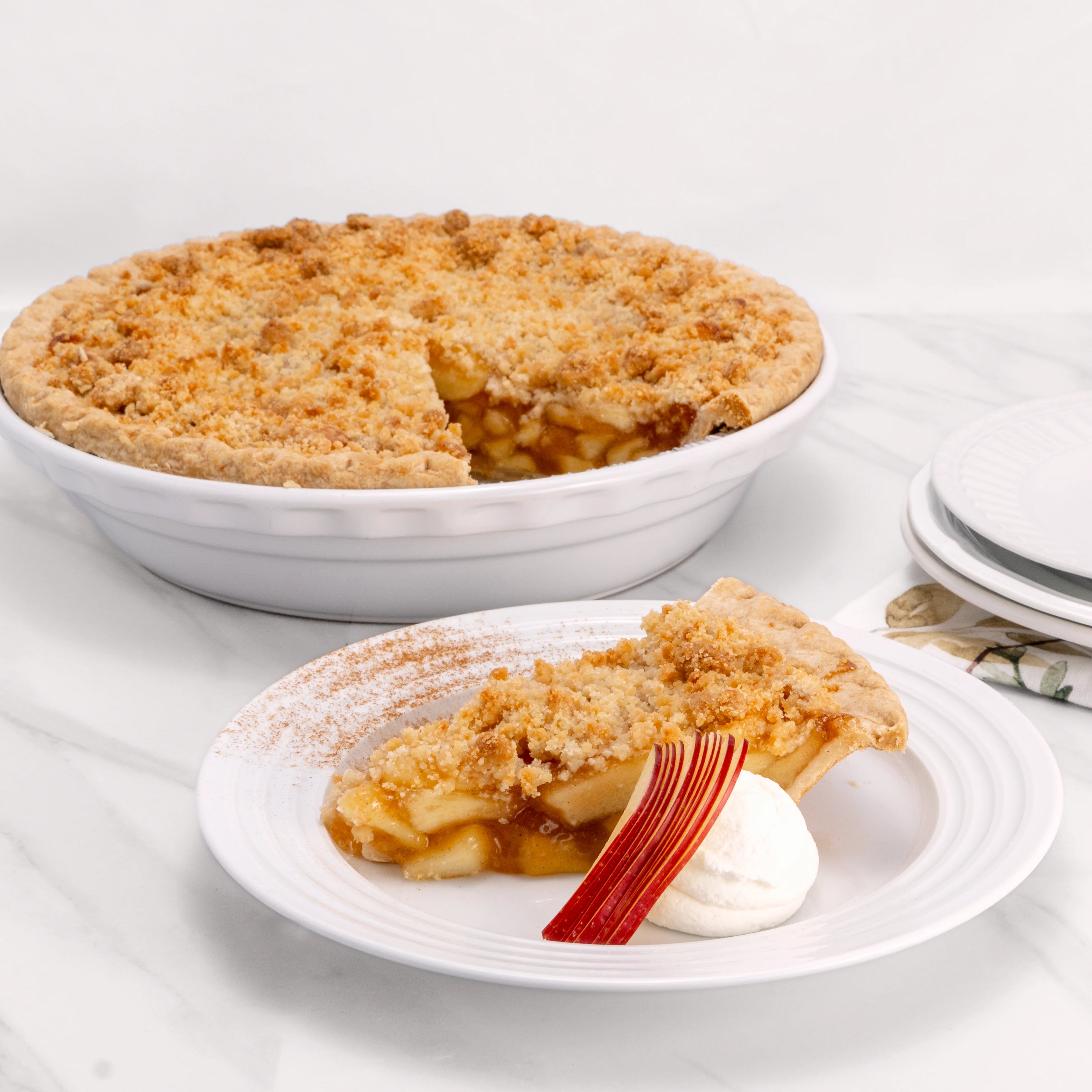 Slice of Apple Crumb Pie, garnished with cinnamon, sliced cheese, and a dollop of crème, in front of an 11" pie from which it was cut.