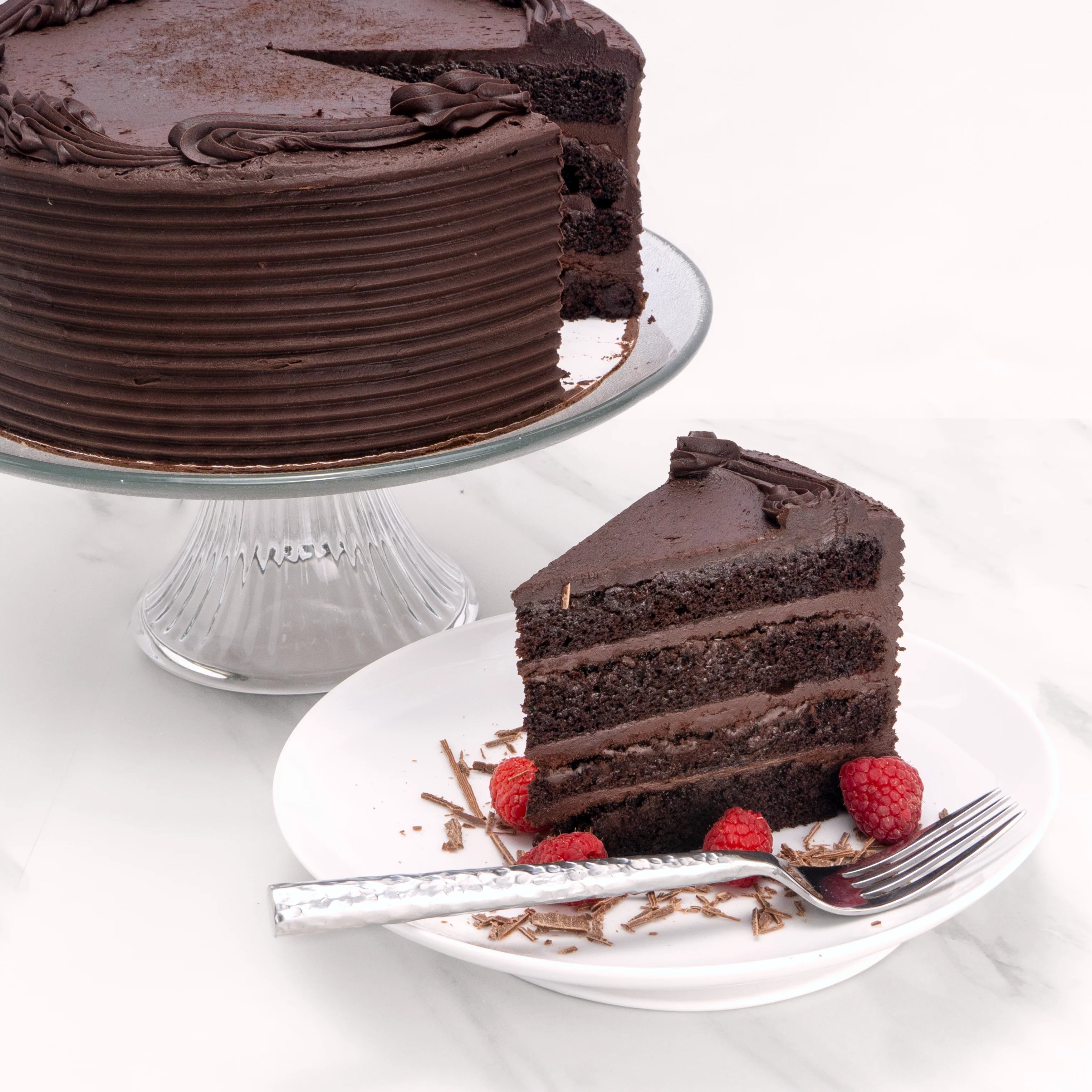 Slice of Heavenly Chocolate Cake, garnished with raspberries and chocolate, in front of a 9" cake from which it was cut.