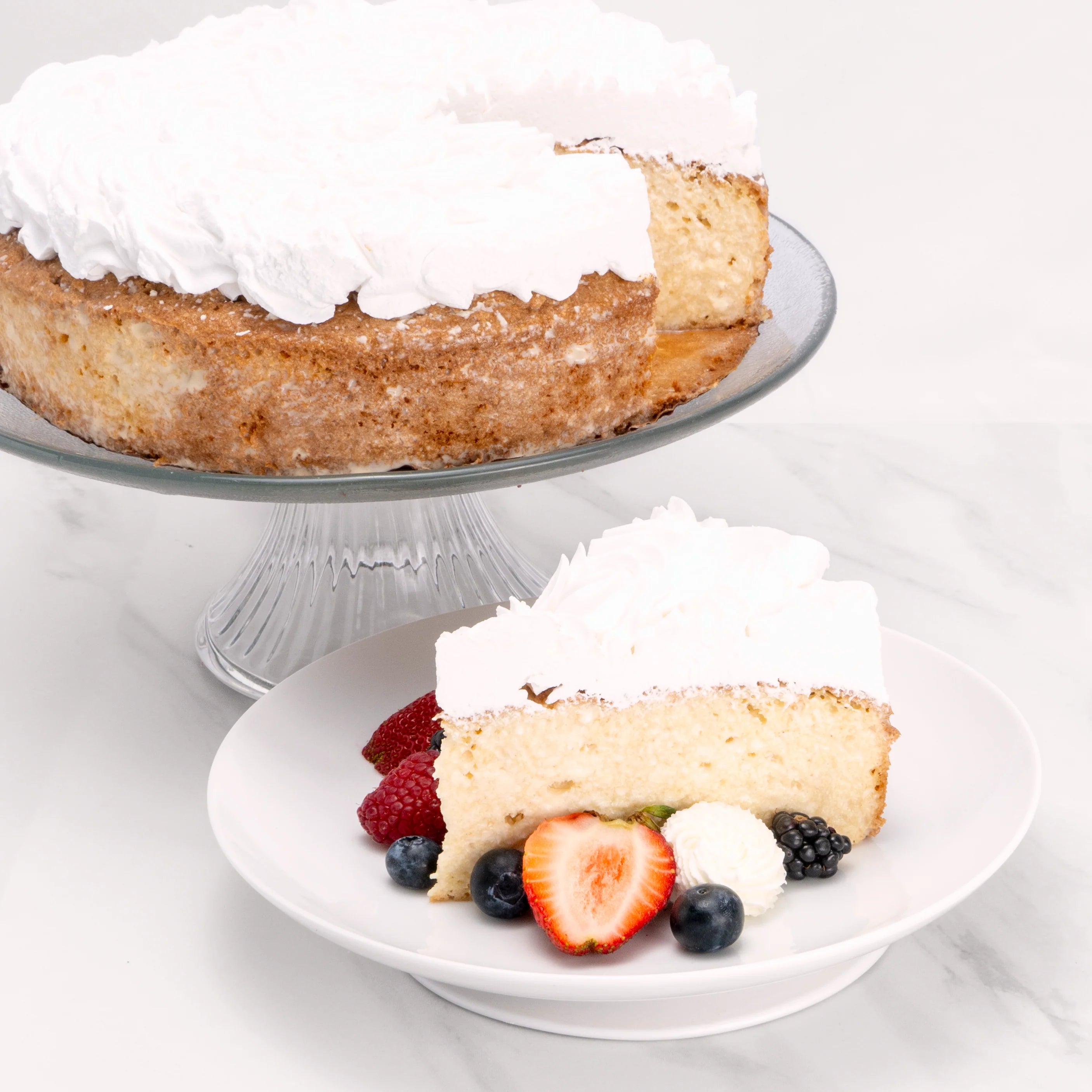 Slice of Tres Leches Cake, garnished with blueberries, strawberries, blackberries, and crème, in front of a 9" cake from which it was cut.