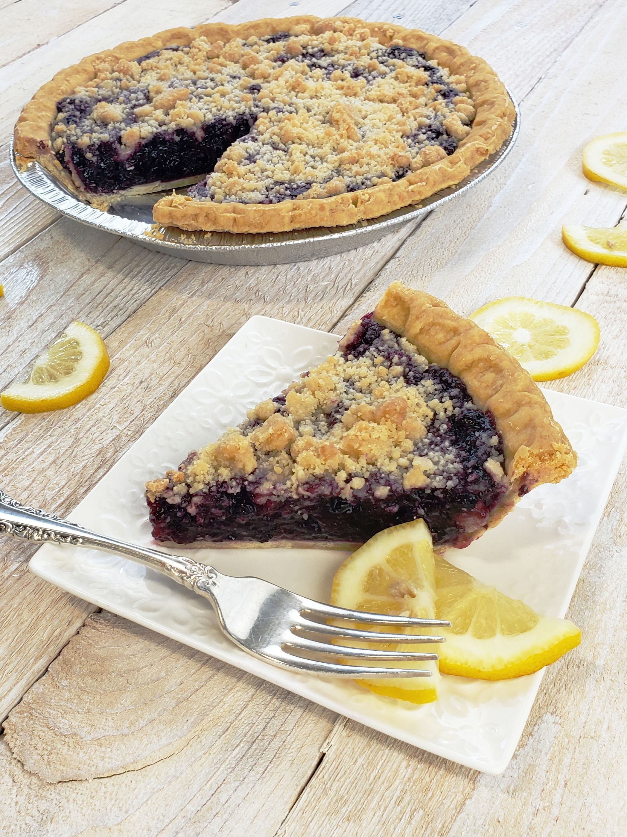 Slice of Blueberry Lemon Vodka pie, garnished with lemon slices and a crumb topping, in front of an 8" pie from which it was cut.