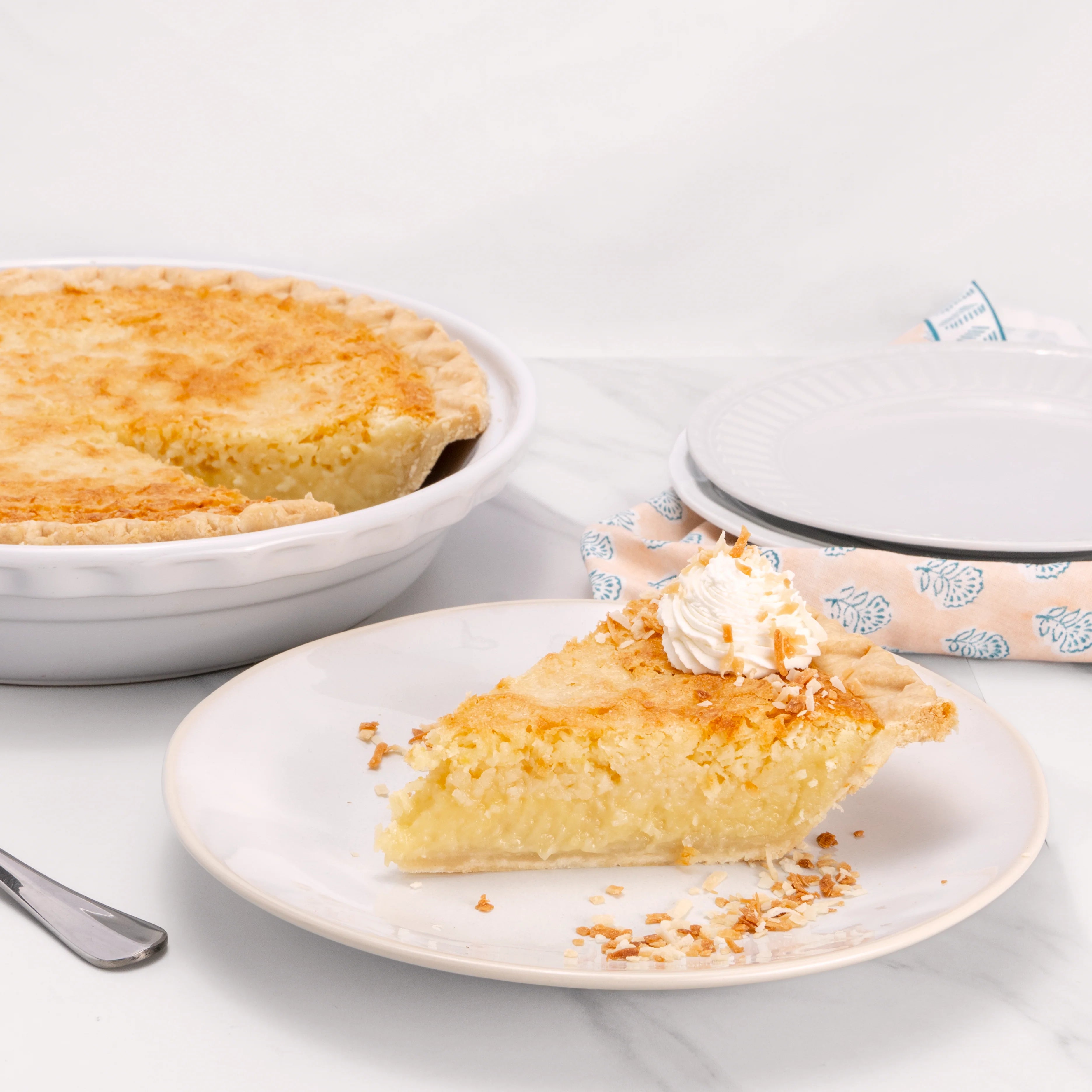 Slice of Coconut Supreme pie, garnished with a dollop of crème on top and toasted coconut flakes, in front of an 11" pie from which it was cut.