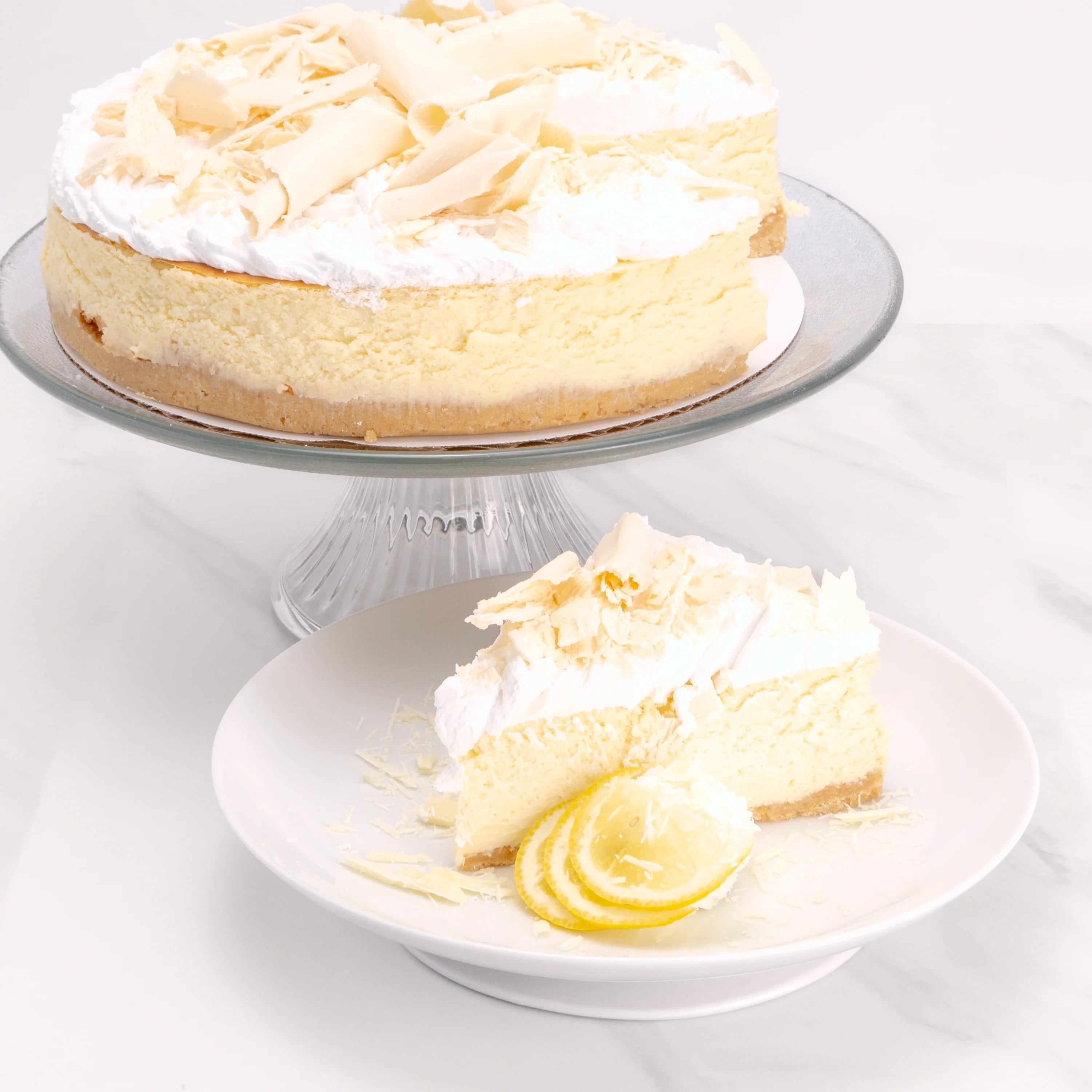 Slice of Gluten Free White Chocolate Crème Cake, garnished with lime, white chocolate, and a dollop of crème, in front of a 9" cake from which it was cut.