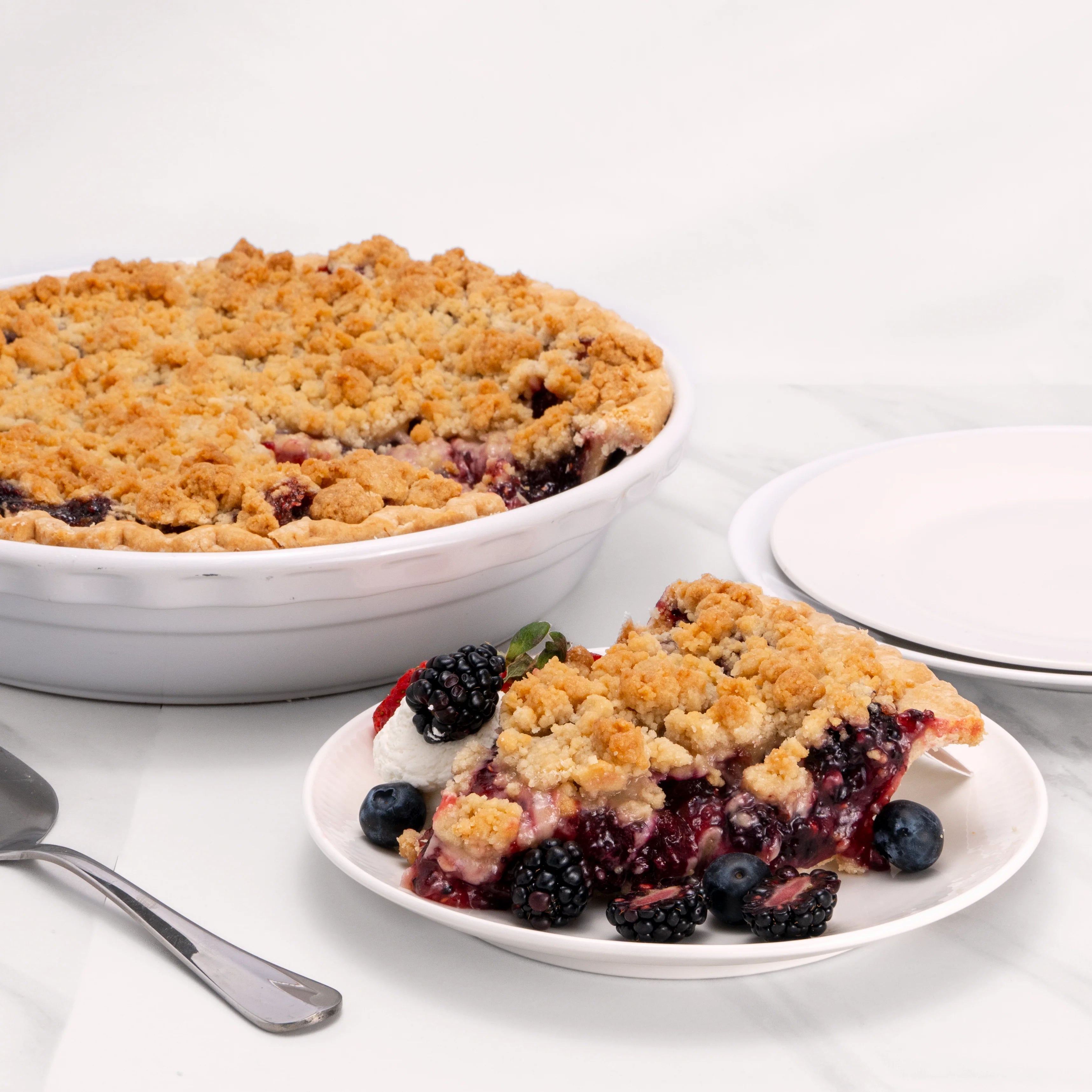 Slice of Triple Berry Pie, garnished with blueberries, strawberries, blackberries, and crème, in front of an 11" pie from which it was cut.