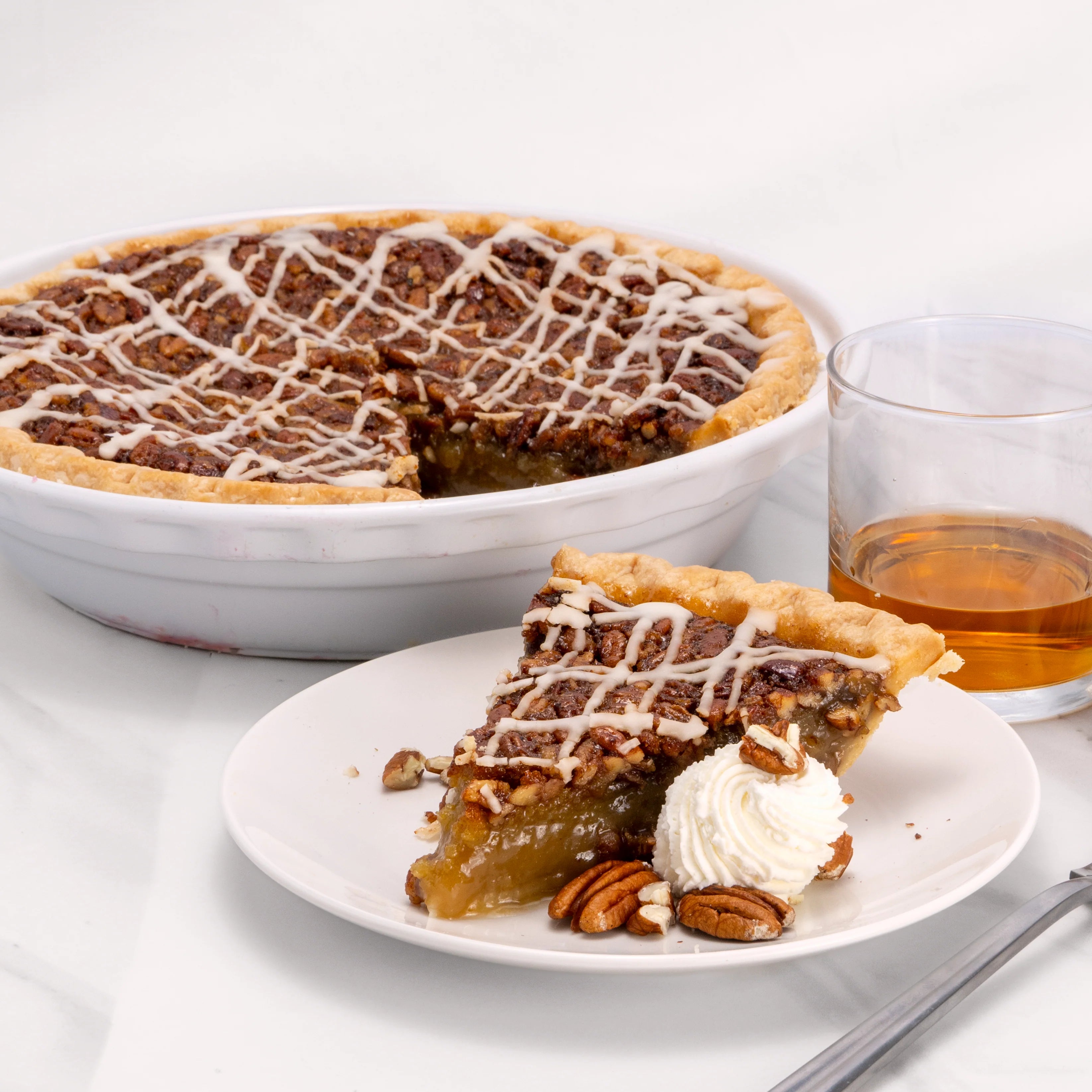 Slice of Whiskey Pecan Pie, garnished with pecans and creme, in front of an 11" pie from which it was cut and a glass of whiskey.